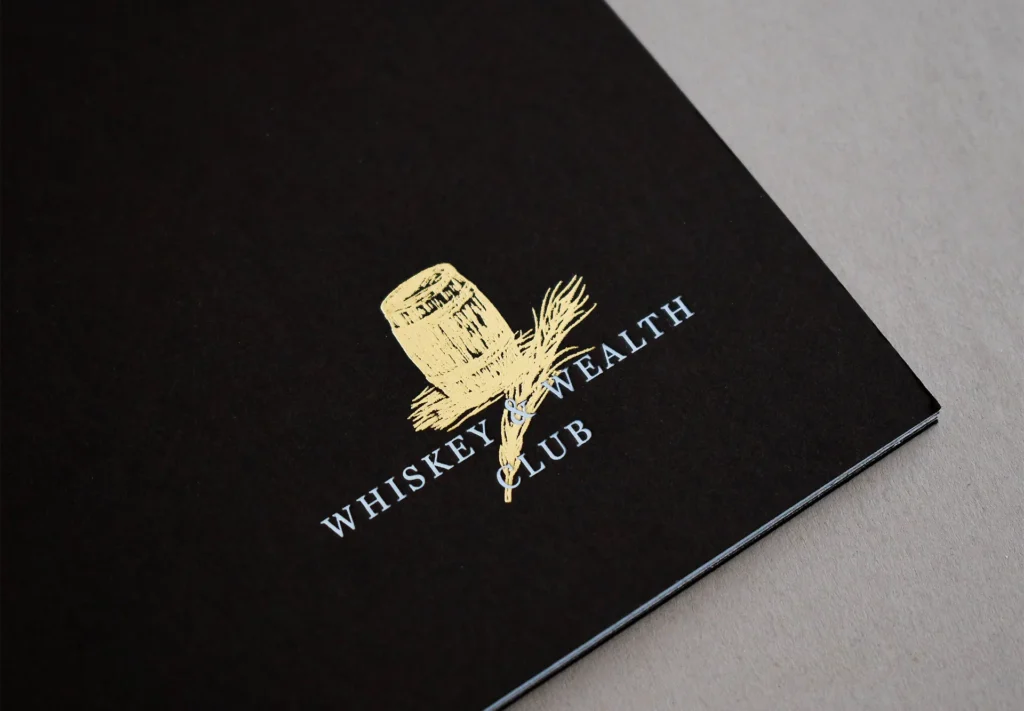 A close-up view showcasing the gold foiling on the logo of a Whiskey & Wealth Club brochure, adding a touch of luxury and sophistication.