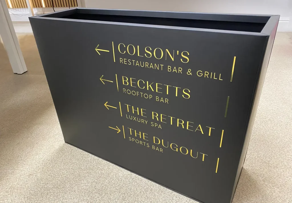 Large, stylish plant box featuring gold vinyl lettering and directional arrows, designed to guide visitors to a specific destination while adding a touch of modern elegance.